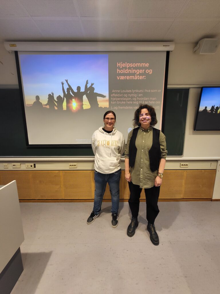 Bilde av to kvinner som står foran et lerret På lerretet er det er det bilde av en solnedgang og siluetter av flere folk, samt teksten "Hjelpsomme holdninger og væremåter" 