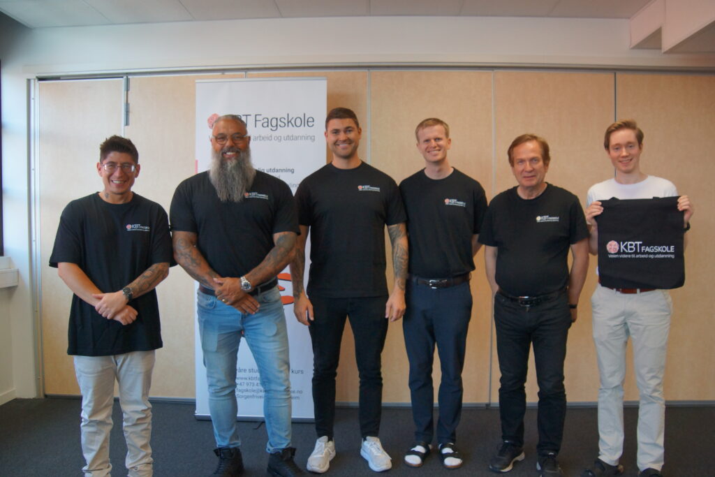 Bilde av representanter fra KBT Fagskole og NextGen Ambassadors. Seks smilende menn står oppstilt foran en rollup med logoen til KBT Fagskole. Mennene har også på seg sorte t-skjorter med logoen til KBT Fagskole på. 