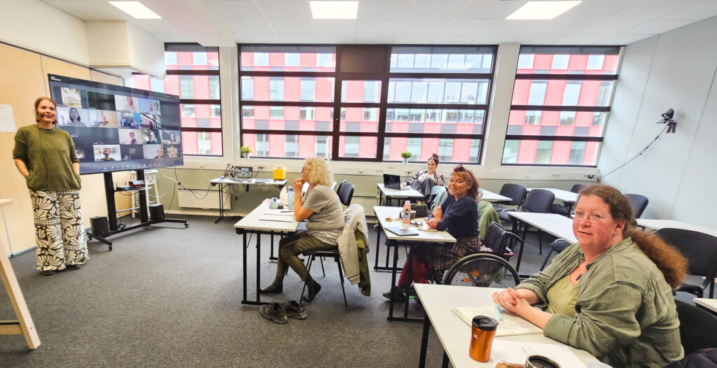 Bilde fra undervisningrom. 4 kvinnelige studenter sitter i rommet, på en skjerm i bakgrunnen ser vi flere studenter. Lærer Sara står ved siden av skjermen. 