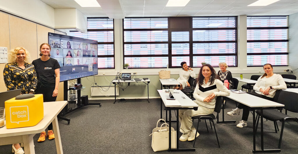 Bilde fra undervisningsrom i Trondheim. På en skjerm i hjørnet ser vi studentene som deltar digitalt. I rommet står lærer Sara og studiekonsulent Olga. Fire kvinnelige studenter sitter ved pulter og ser mot kamera. 