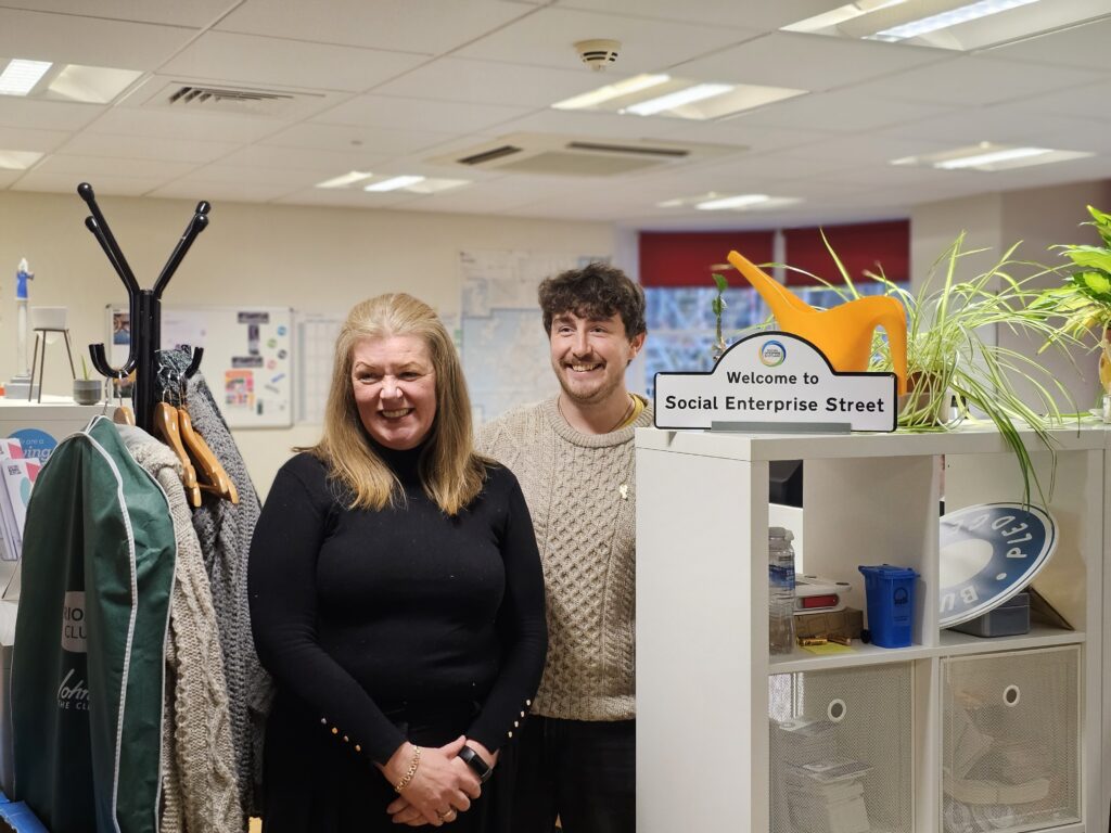 Bilde av en smilende dame i sort genser og en smilende ung mann i beige genser. De står mellom en stumtjener og en bokhylle. På bokhyllen er det et skilt: Welcome to Social Enterprise Street 