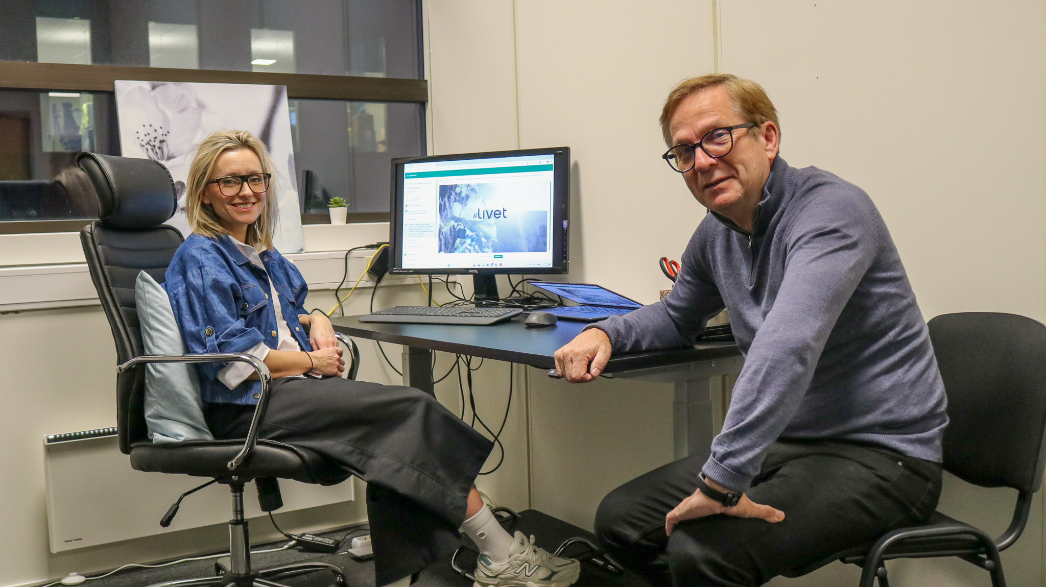 Bilde av en kvinne og en mann som sitter ved en skriveput inne på et kontor. På dataskjermen i bakgrunnen står det #Livet