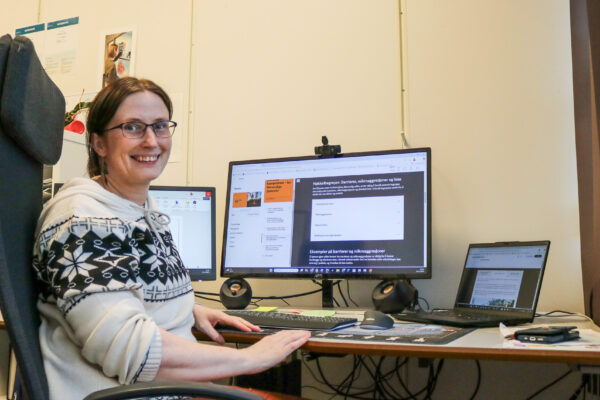 Bilde av Yvonne Vornes. Hun sitter ved et skrivebord og smiler mot kamera. I bakgrunnen er dataskjermer, med utsnitt fra et e-læringskurs