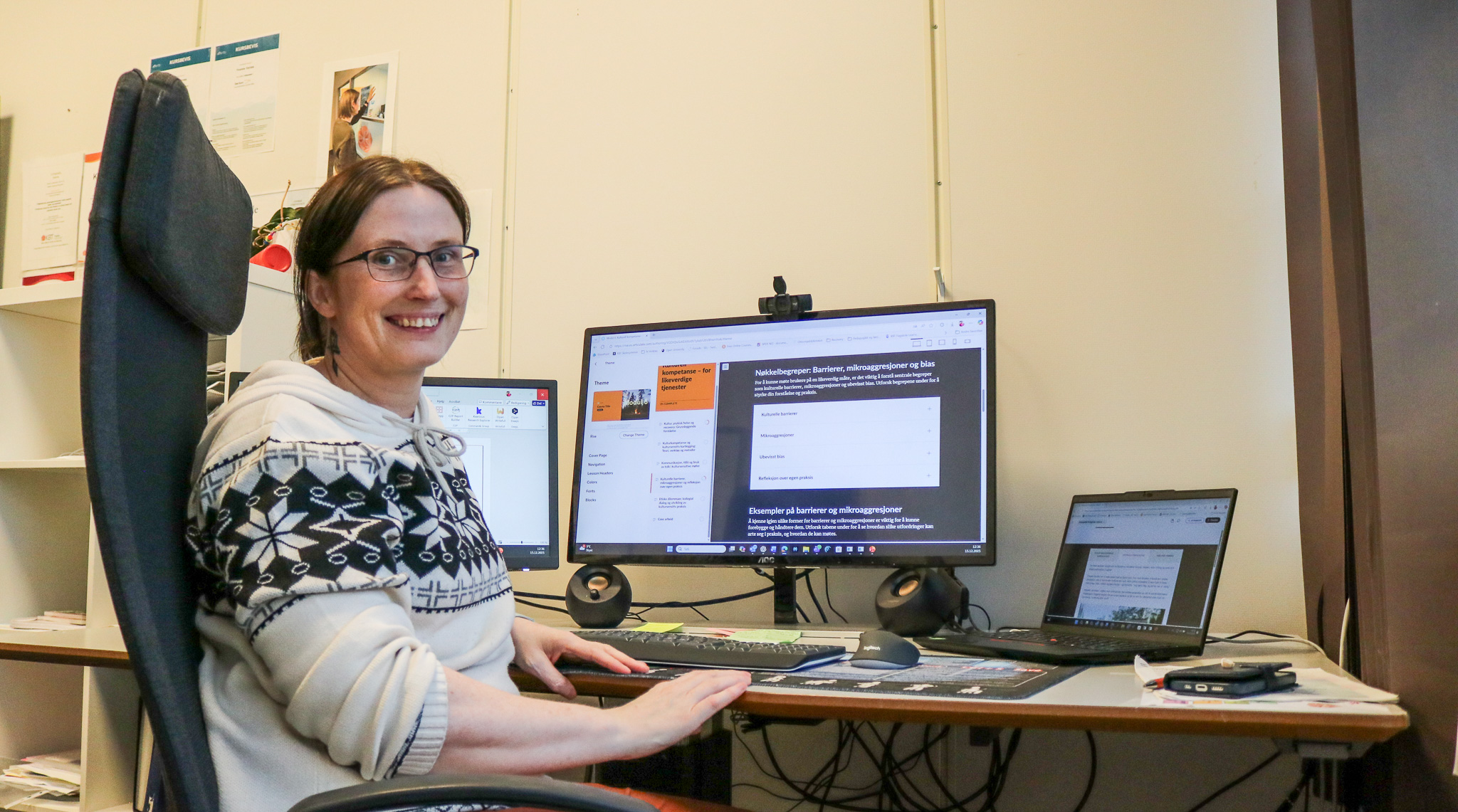 Bilde av Yvonne Vornes. Hun sitter ved et skrivebord og smiler mot kamera. I bakgrunnen er dataskjermer, med utsnitt fra et e-læringskurs