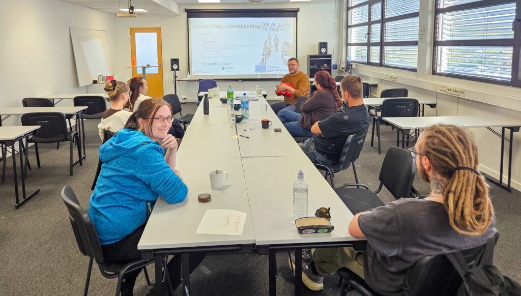 Bilde av flere personer som sitter rundt et langt bord. En mann holder en rød boks. En kvinne i blå genser ser mot kamera og smiler. 