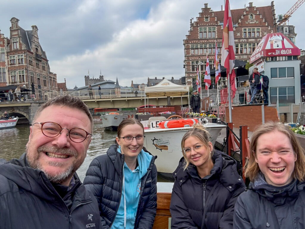 Bilde av fire smilende personen. I bakgrunnen ser vi bygninger fra en Europeisk by 