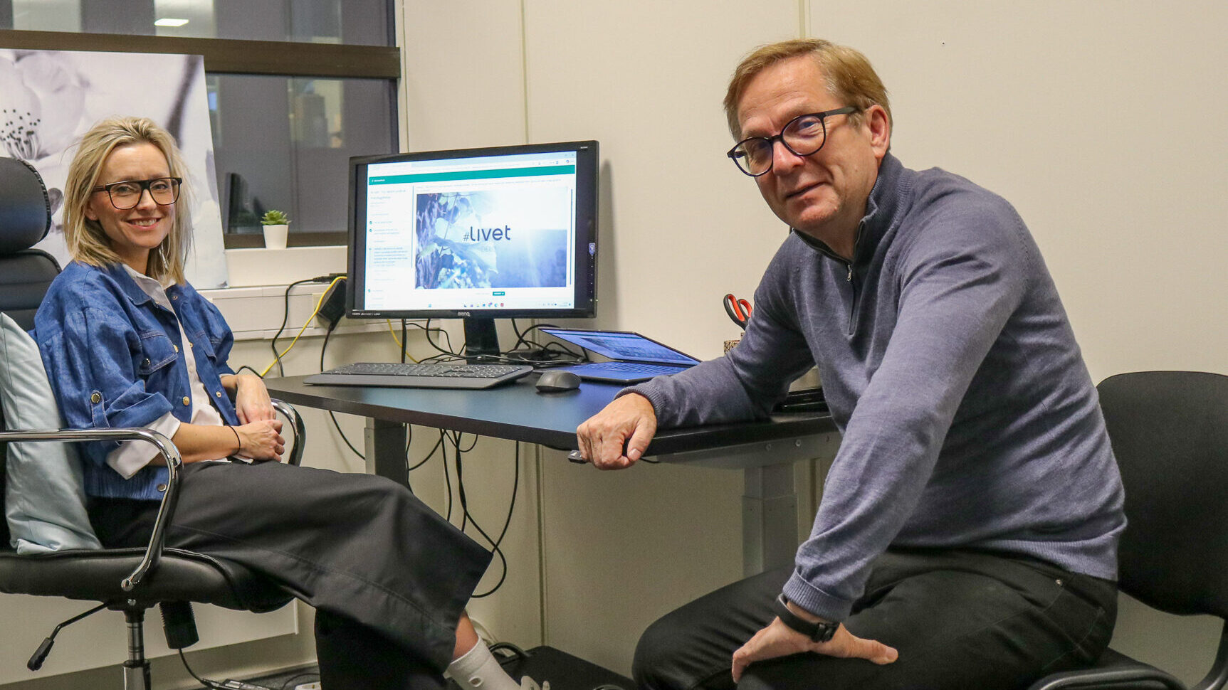 Bilde av en kvinne og en mann som sitter ved en skriveput inne på et kontor. På dataskjermen i bakgrunnen står det #Livet