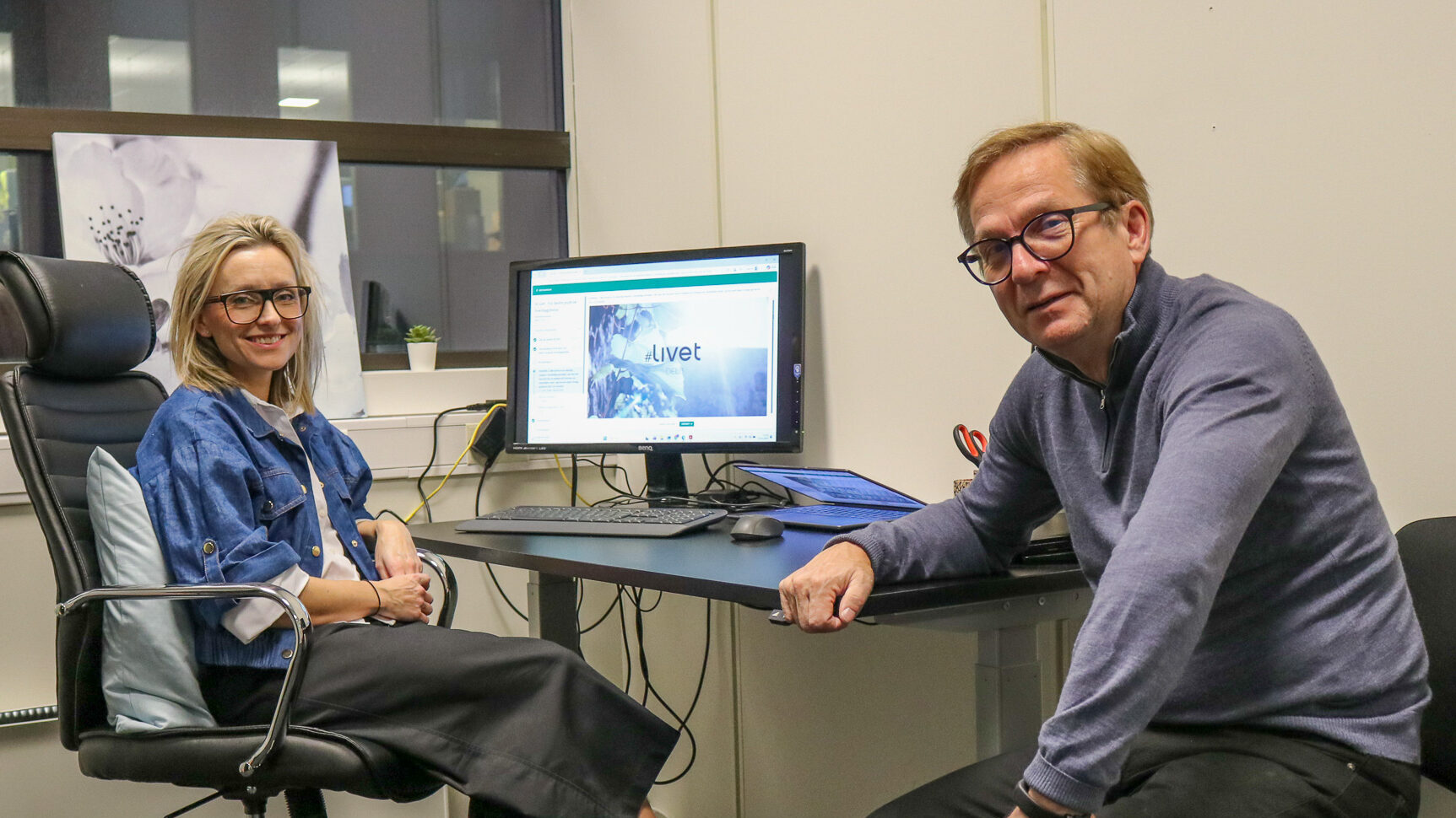 Bilde av en kvinne og en mann som sitter ved en skriveput inne på et kontor. På dataskjermen i bakgrunnen står det #Livet