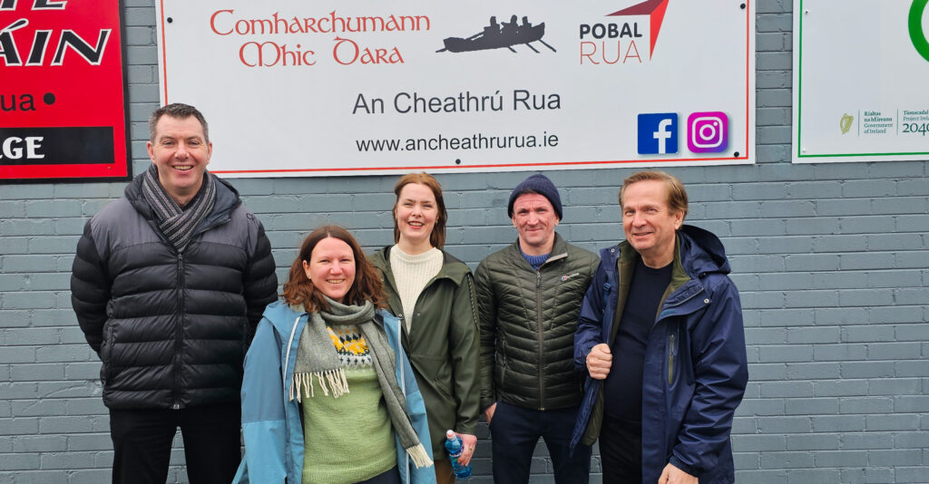 Bilde av tre menn og to kvinner som står foran en vegg. På veggen er det plakater med irsk tekst. 