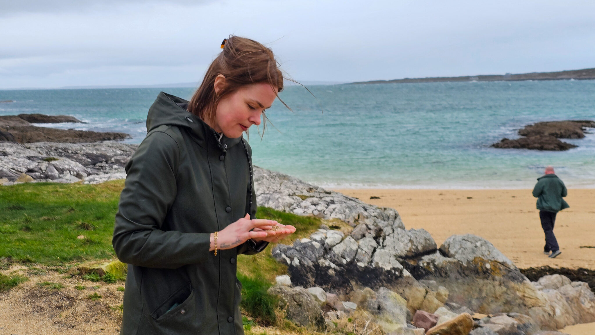 Bilde av en kvinne i regntøy som står på en strand. I hånden har hun sand. 