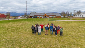 A group of people standing in front of a yellow builing, the surroundings are rural with farms and a big lawn