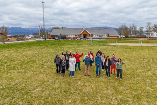 A group of people standing in front of a yellow builing, the surroundings are rural with farms and a big lawn